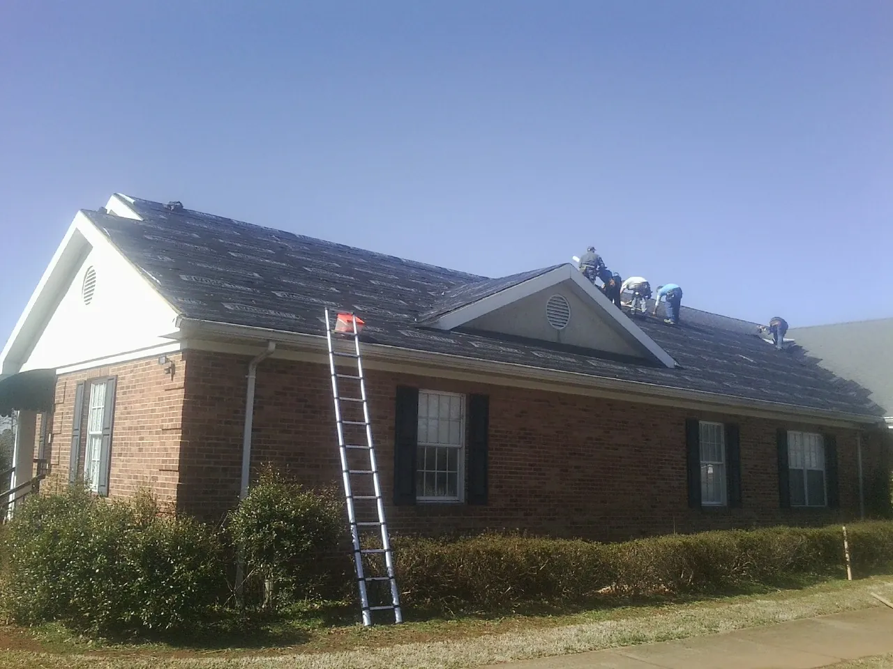 Crew tearing shingles off a brick ranch roof with a ladder set — Emergency Roof Repair work by our crew in Rome