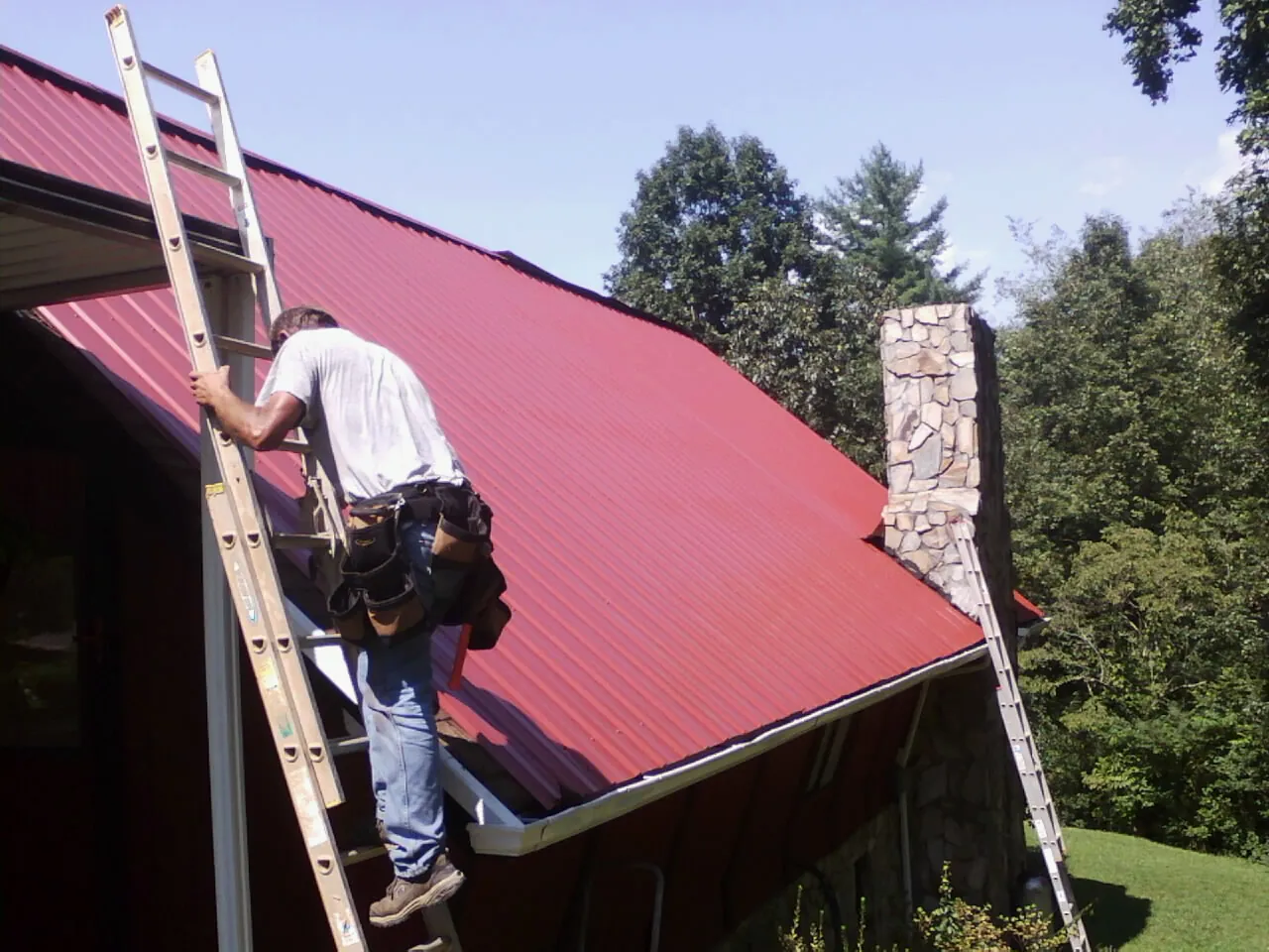 Roofer on a ladder beside a red metal roof and stone chimney — Soffit & Fascia Repair by Roofing Brothers in Rome